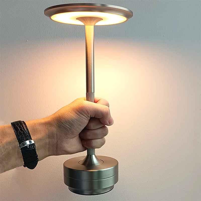 Hand holding a modern silver portable LED table lamp against a plain wall.
