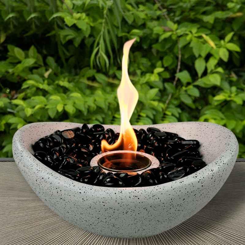 Tabletop fire bowl with black stones on outdoor patio, flame burning, green foliage background