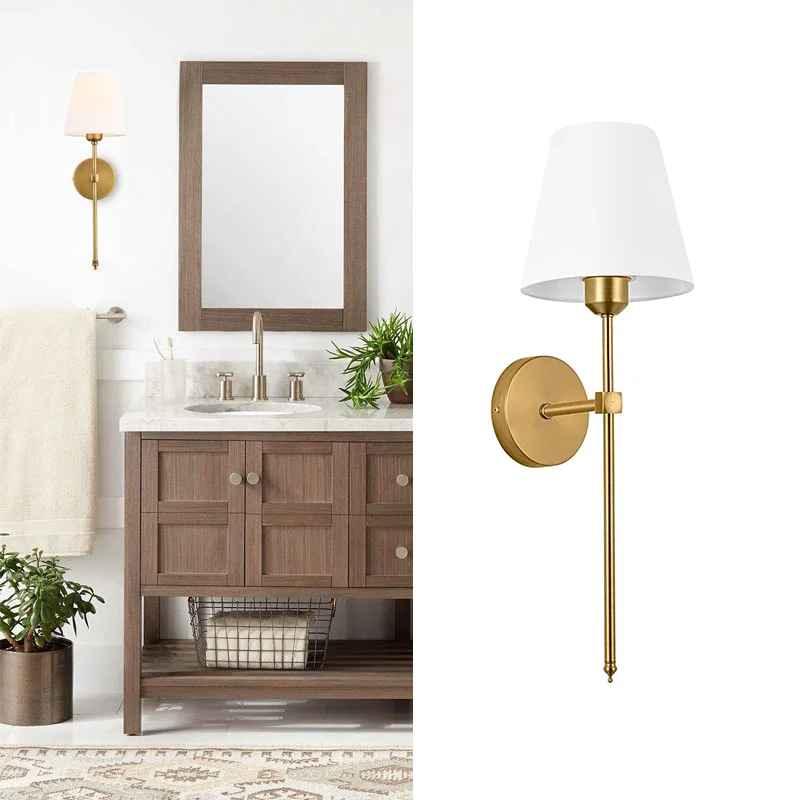 Modern brass wall sconce with white shade in a stylish bathroom with wooden vanity and plants